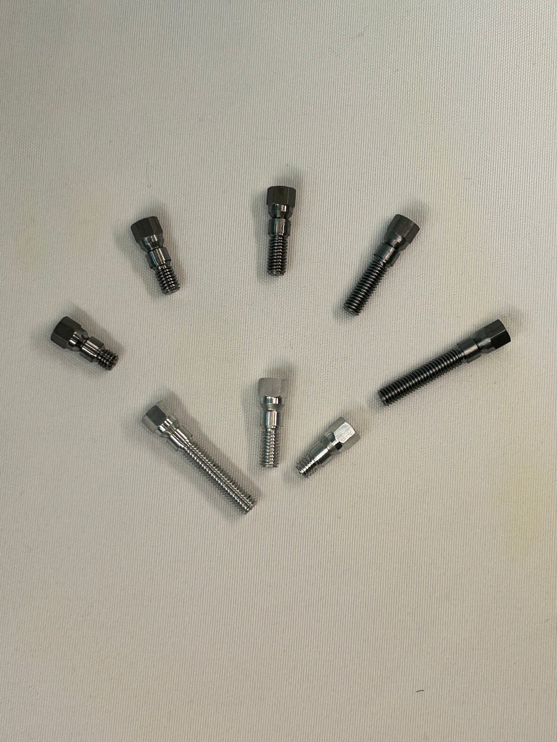 Seven dark metallic threaded hardware components arranged in a loose circle on a plain off-white surface. Size 2 stitching Pins, Steel and Aluminum. 