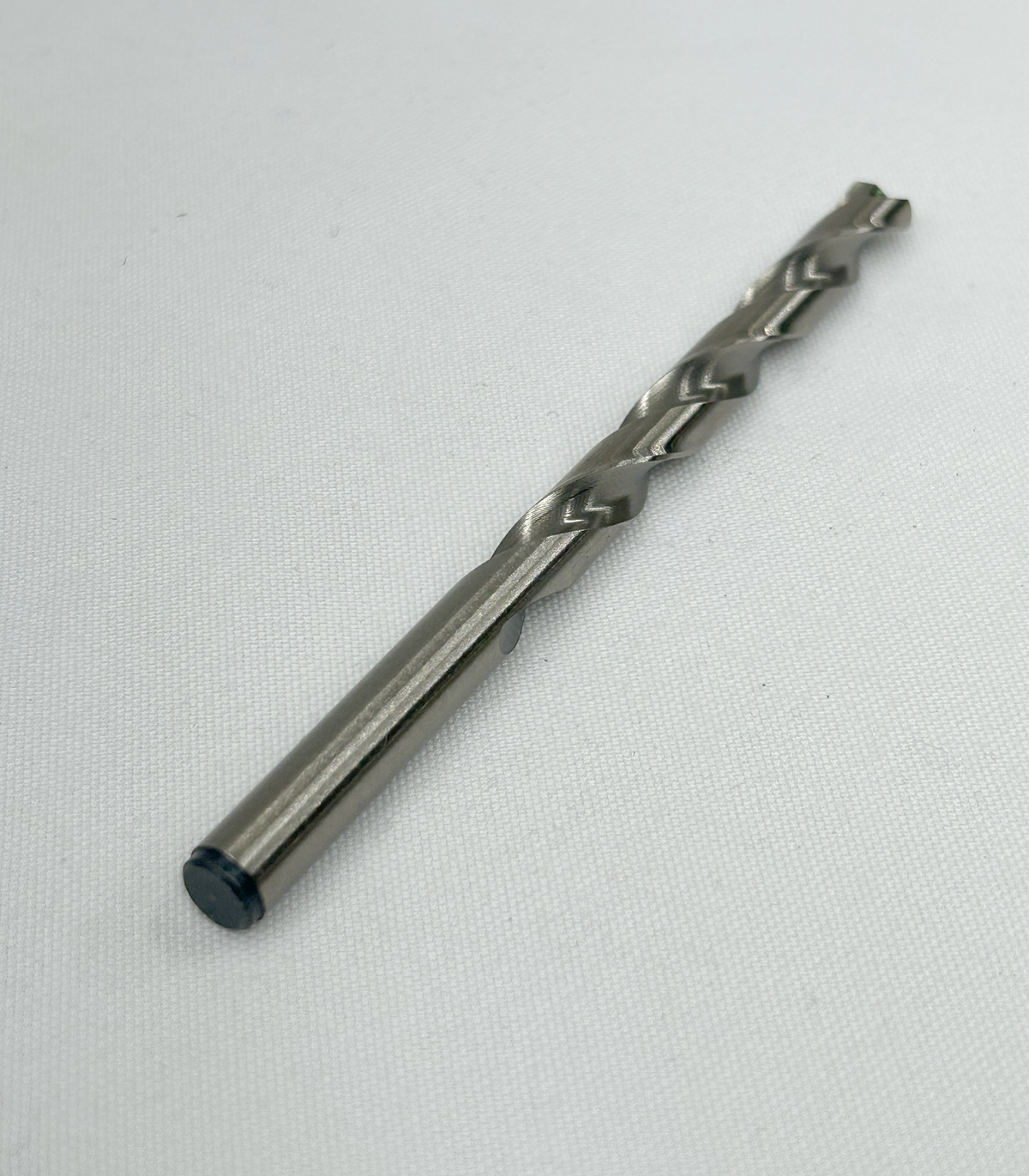 A single metal drill bit with a dark protective tip, photographed diagonally on a textured white fabric background.