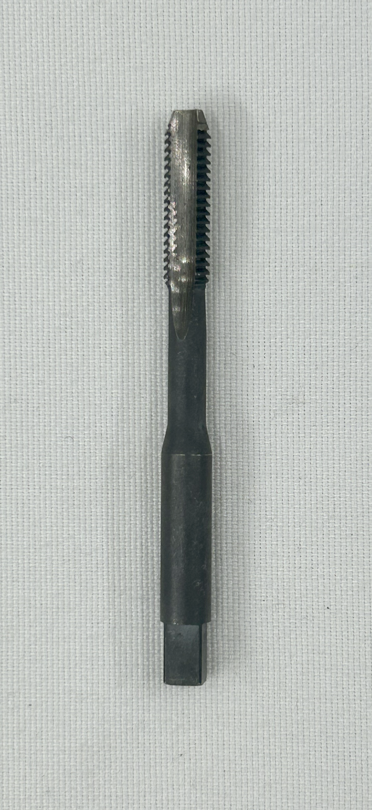A black metal thread cutting tool with a threaded cutting head and a square socket at the end, photographed on a light gray textured fabric background.