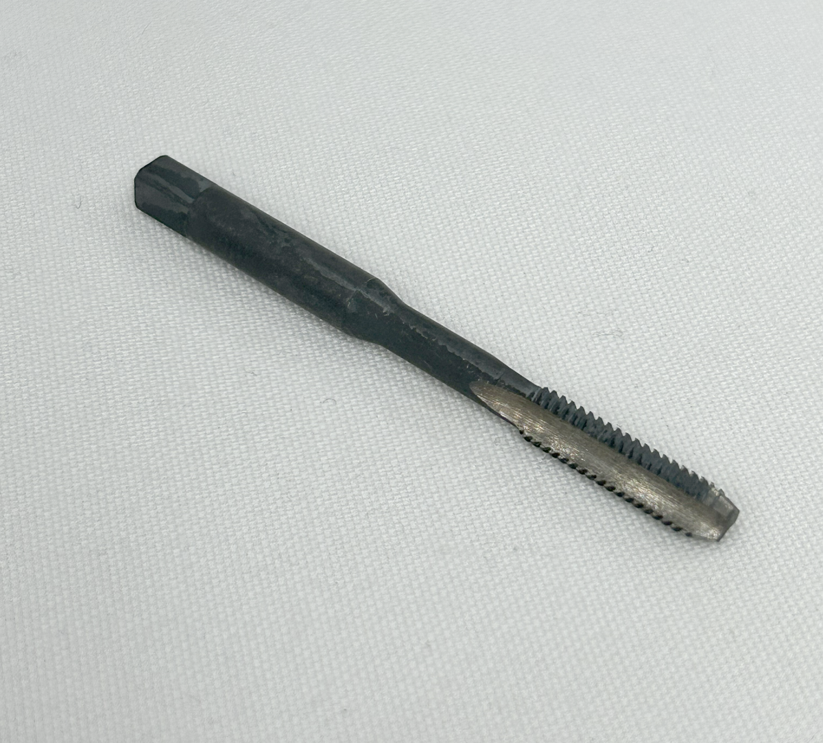 A black metal thread cutting tool with a threaded cutting head and a square socket at the end, photographed on a light gray textured fabric background.