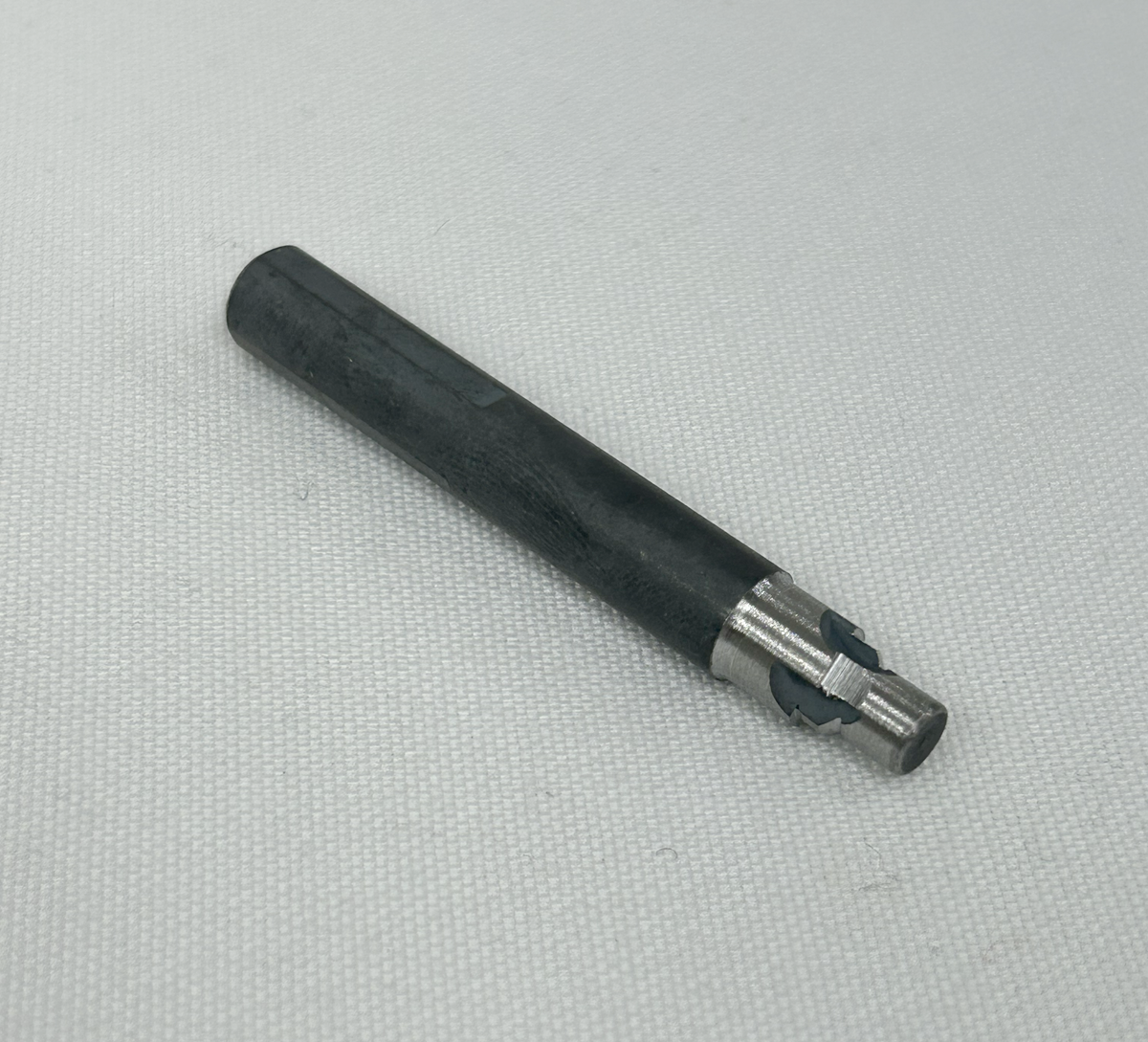 A small cylindrical metal tool bit with a black coated body and a silver exposed cutting section, photographed diagonally on a white textured fabric background.