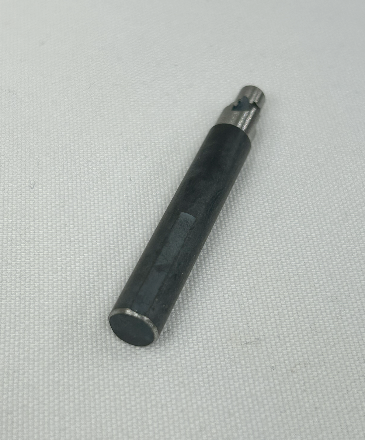 A black cylindrical metal tool or pen-like accessory with a silver tip, placed diagonally on a textured white fabric background.