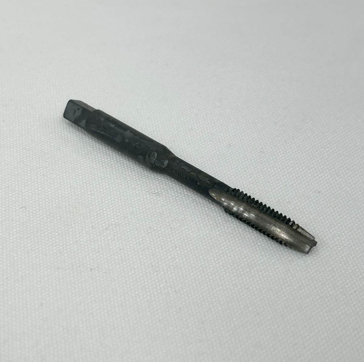 A single dark metal threaded tool bit, likely a tap or reamer, placed diagonally on a textured white fabric background.