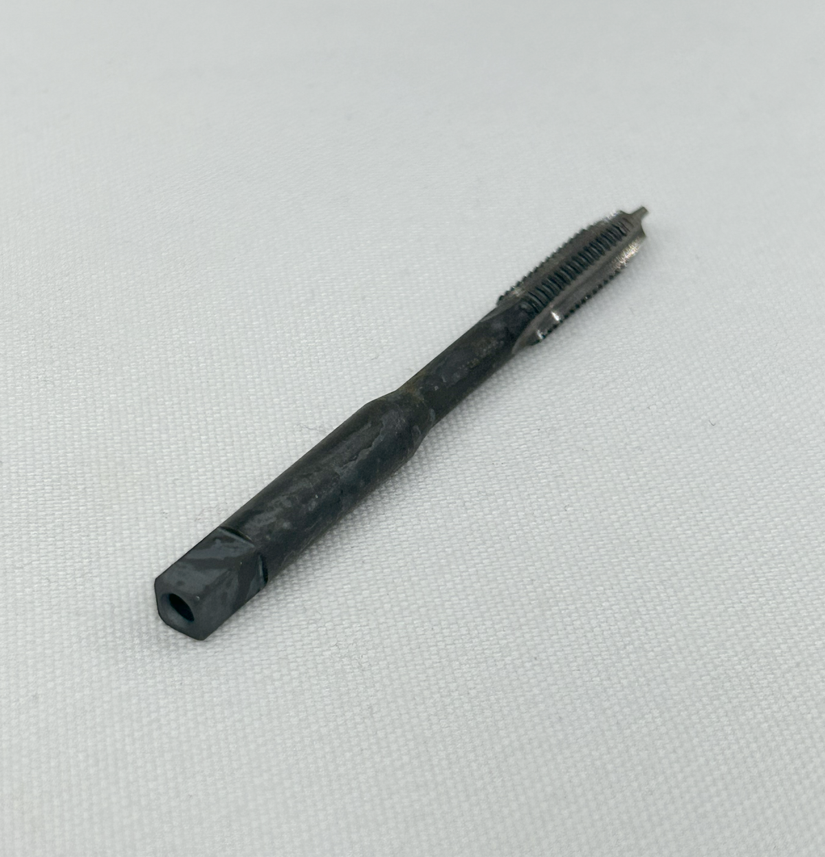 A black metal threading tool bit, likely a tap or reamer, resting diagonally on a textured white fabric background.